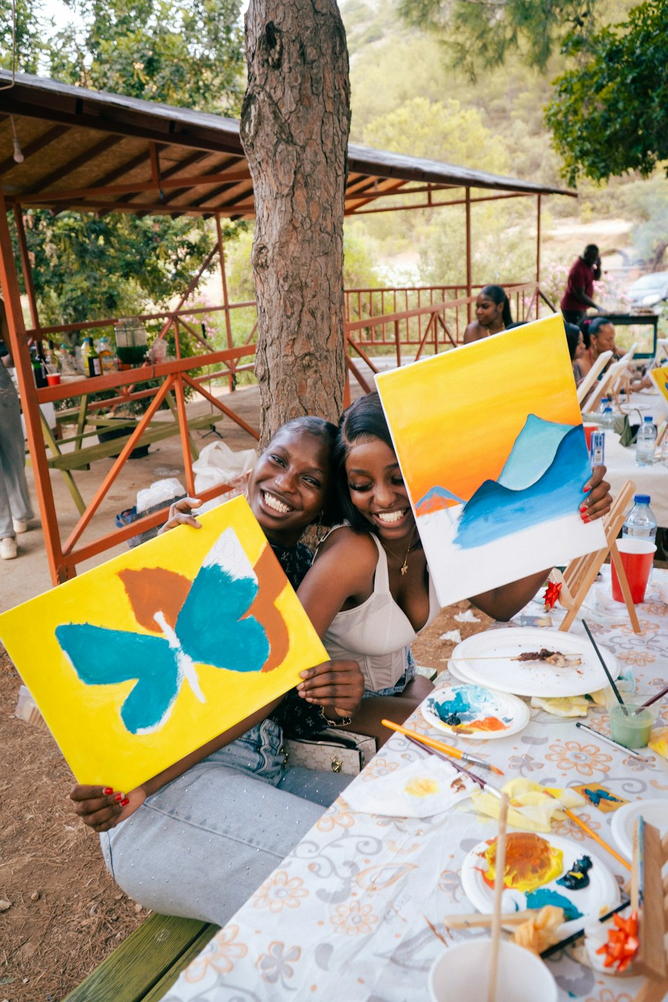Two smiling women show off their paintings at an outdoor art class.